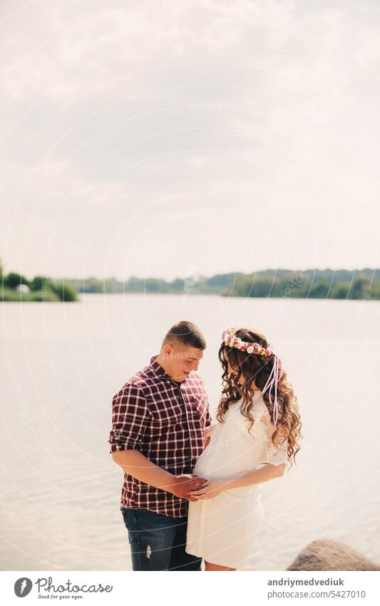 Young happy romantic pregnant couple hugging on nature near lake in summer park. Pregnant woman expecting a baby. Future mom and dad, family. mother's, father's day