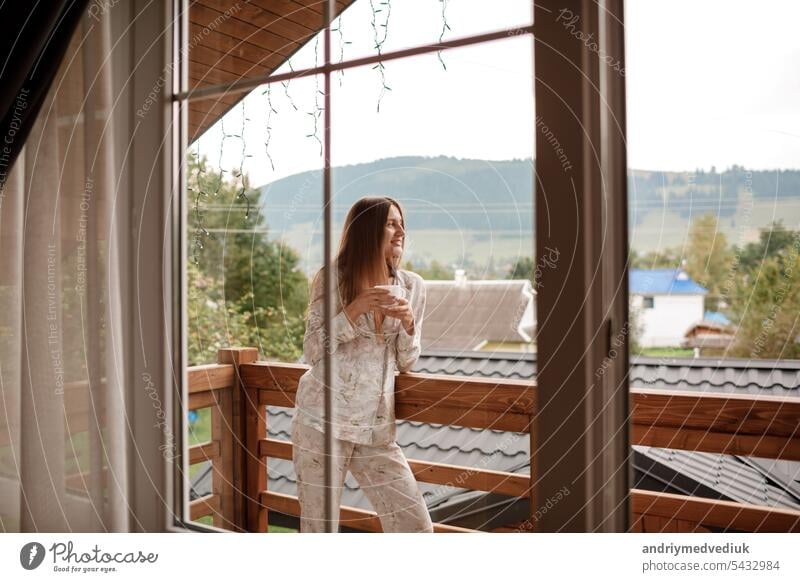 Young female standing after taking a shower in the morning on balcony of the hotel. holding a cup of coffee or tea in her hands. Looking outside nature forest and Mountain