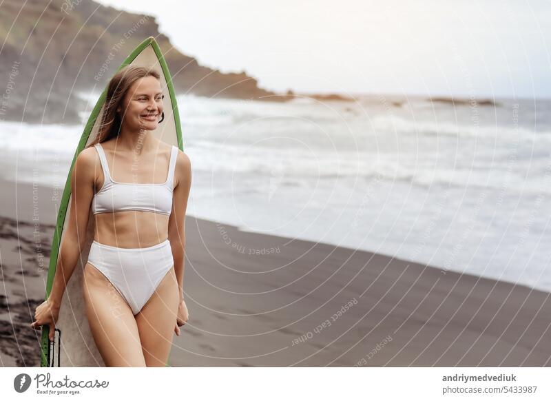 Young surfer woman in bikini going surfing stands with surfboard on the black sandy beach. Girl walks with longboard. Extreme water sport by the ocean, sea. Healthy Active Lifestyle. Summer Vacation