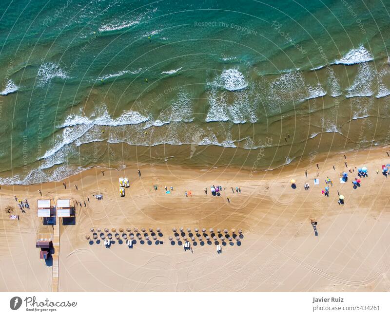 zenithal aerial view of a beach in summer with bathers, water skates and thatched beach umbrellas for rent, handicapped access ramp and lookout tower seen from above, drone view