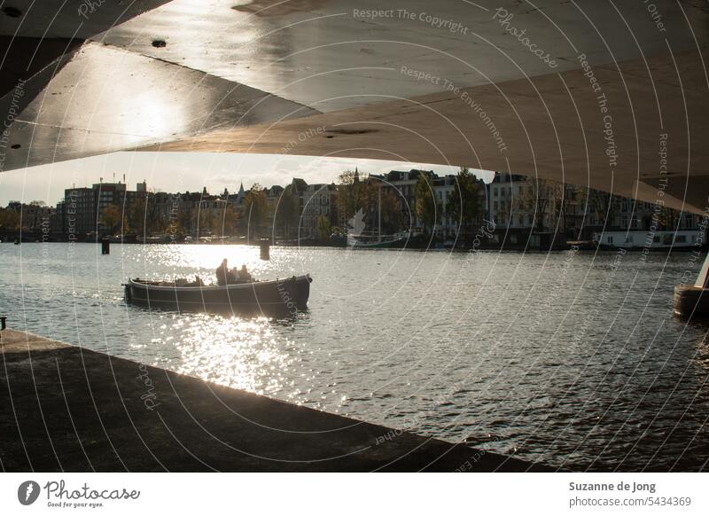 boat in the sun photographed under a bridge along the Amsterdam canals. The sun is shining which results in a silhouette of the boat. The bridge also catches sun and is shiny. The image has an idyllic, chill and urban vibe.