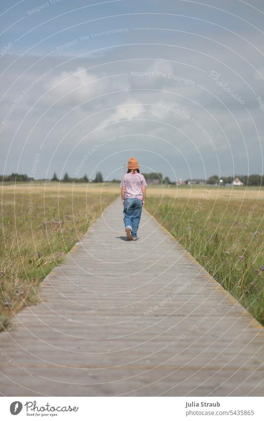 Girl walking on a footbridge Footbridge Salt meadow North Sea Easygoing Cap hands bags Jeans Summer warm wooden walkway long way Forward cloudy sky Grass Green