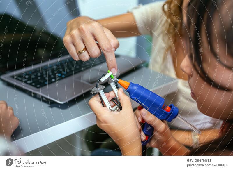 Teacher helping to student using hot melt glue gun on machine pieces in robotics class unrecognizable teacher female assist support kid children electronic