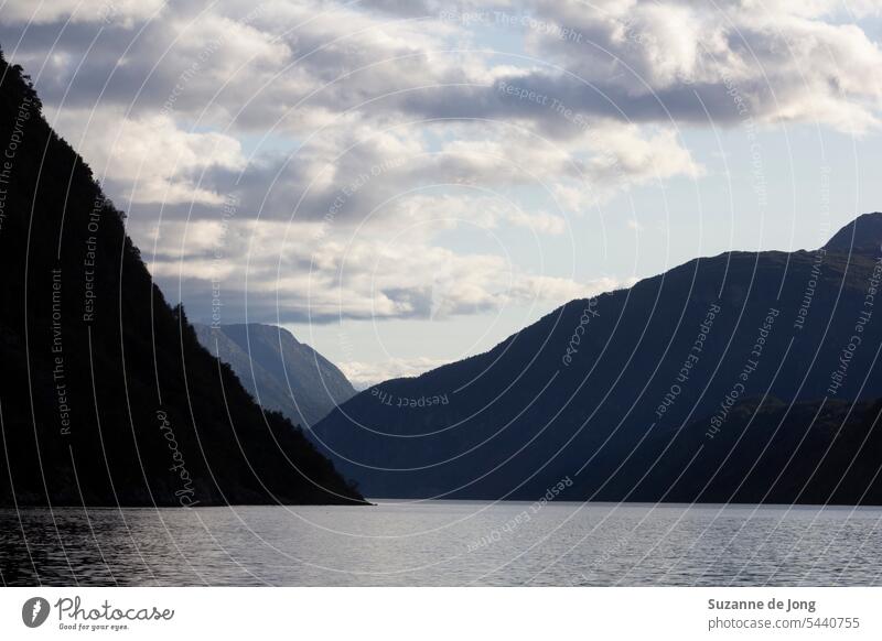 Picturesque view on a fjord and mountains in Norway on a nice summer afternoon, with a blue sky with some clouds. The light behind the mountain creates a beautiful silhouette. The vibe of the image is calming.