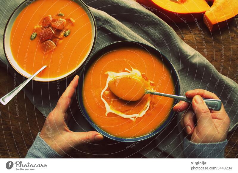 Flat-lay of female hands and Fall warming pumpkin cream soup with croutons and seeds on board over rustic wooden background, top view. Autumn vegetarian, vegan, healthy comfort food eating concep