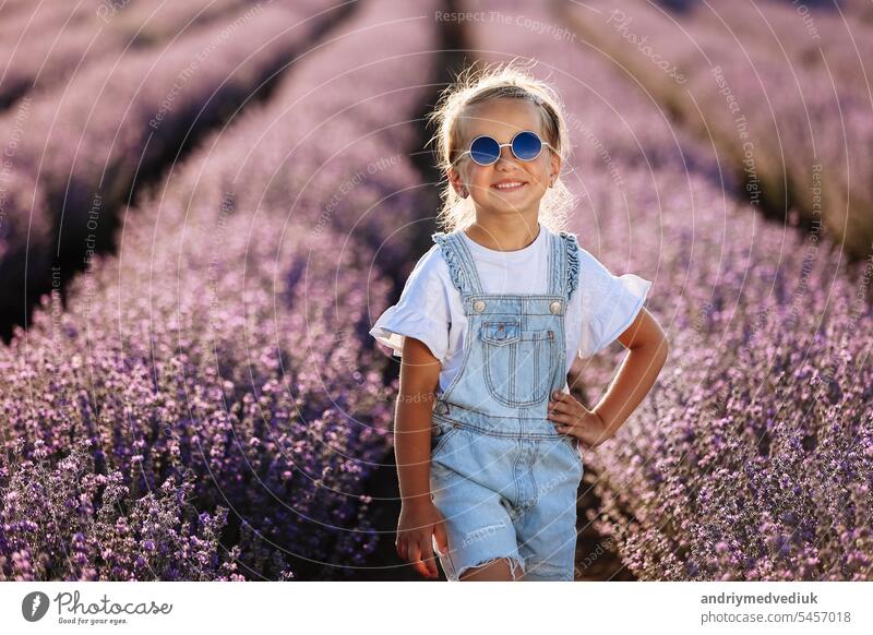 adorable child girl in lavender field on sunset. smiling kid in sunglasses, jeans jumpsuit is having fun on nature on summer day. Family day, vacation, holiday.International Children's Day,