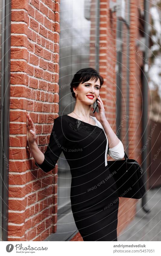 Happy lawyer businesswoman professional is walking outdoors talking on cell smart phone. Caucasian successful female is smiling wearing stylish white-black dress with black bag