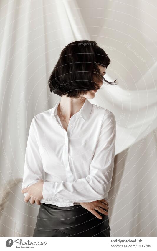 Friendly confident young woman with folded arms smiling at the camera and having fun. Girl in white shirt and leather skirt sitting at home studio attractive