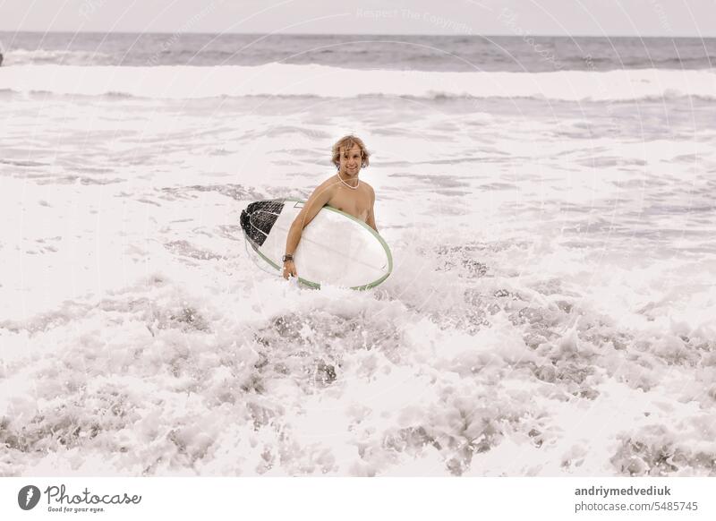 young fit surfer man with surfboard runs into ocean or sea with big waves for surfing. Concept of extreme sport, fitness, freedom, happiness, new modern life. Sports travel destination.