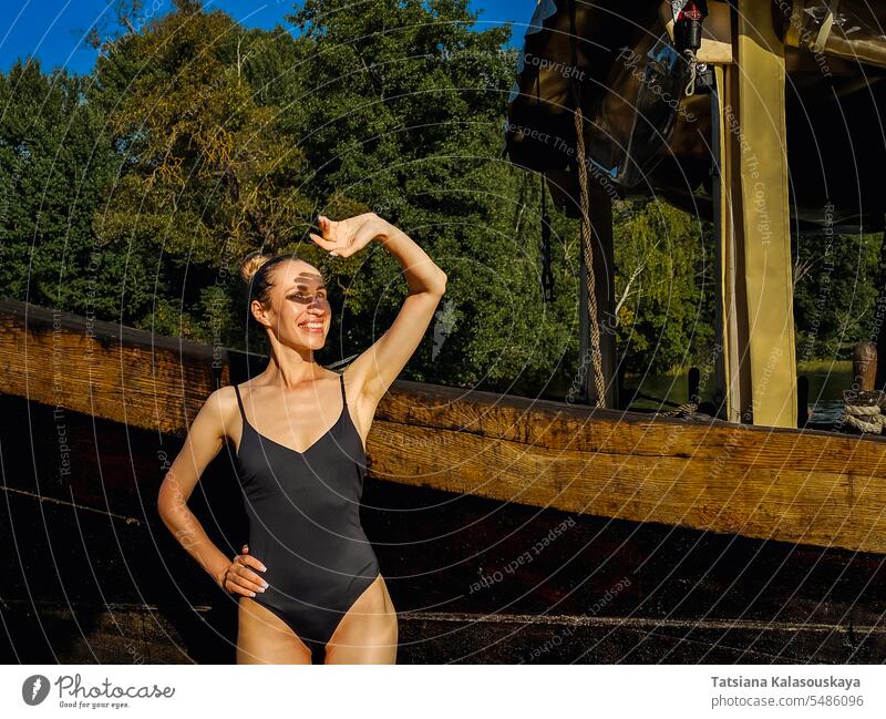 A woman tourist relaxes and smiles, closing her hand from the bright sun while standing near an old wooden ship slim swimsuit water vintage college boat smiling