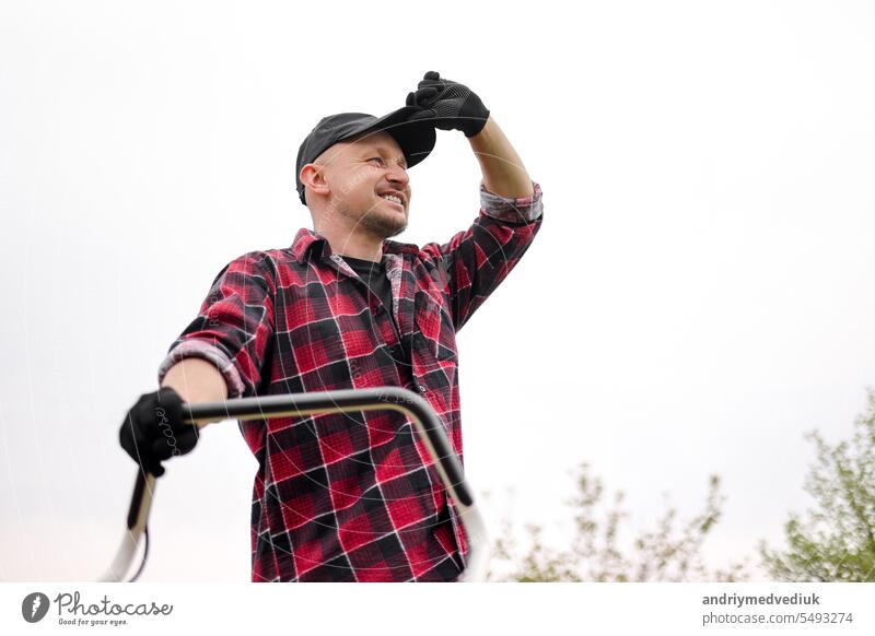 Bottom view of tired man makes a short break while cutting grass with a lawn mower after at backyard of his house. Gardening, sweating or exhausted male resting after working outdoor household chores