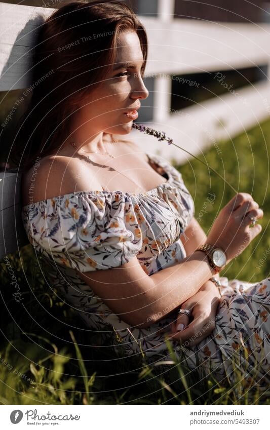 Young beautiful woman in floral sexy dress holds lavender flower in hands sits by the white wooden fence in blooming purple lavender field. Girl enjoying life and dreaming. Natural beauty concept