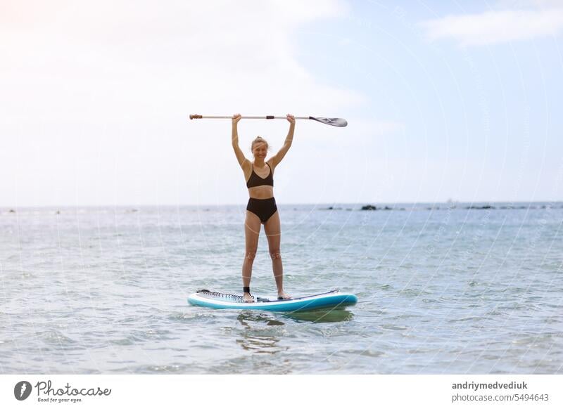 Happy young woman in stylish black bikini raised up the paddle, balancing on a SUP board, on tranquil ocean waters on sunny summer day. Healthy lifestyle and extreme recreation concept.