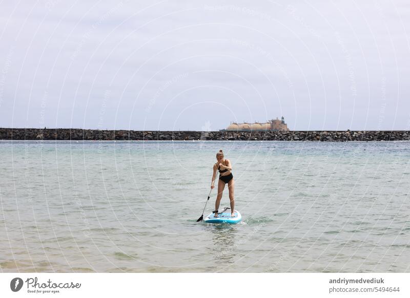 Young woman in stylish black swimsuit with a paddle gracefully balancing on a SUP board, gliding along the tranquil ocean on sunny summer day. Healthy lifestyle and extreme recreation concept.