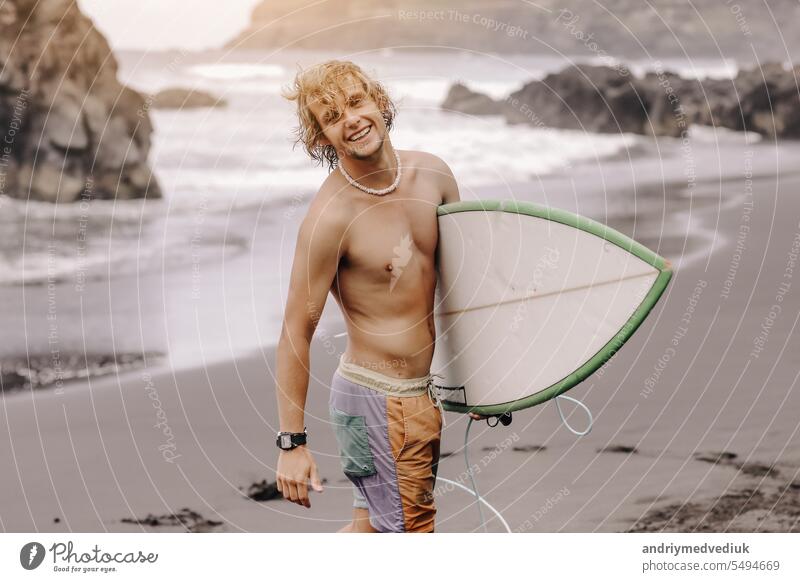 Handsome fit young blond man with mock up surfboard waits for wave to surf spot at sea ocean beach with black sand and looks at camera. Concept of sport, fitness, freedom, happiness, new modern life