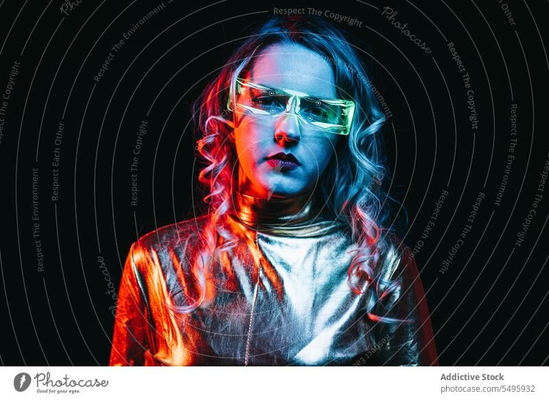 Cybernetic young girl against dark background cybernetic curly hair blonde glasses futuristic wear bright reflecting clothing looking at camera unemotional