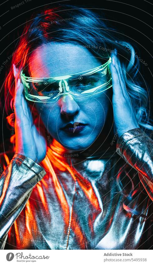 Cybernetic young girl against dark background cybernetic curly hair blonde glasses futuristic wear bright reflecting clothing looking at camera unemotional