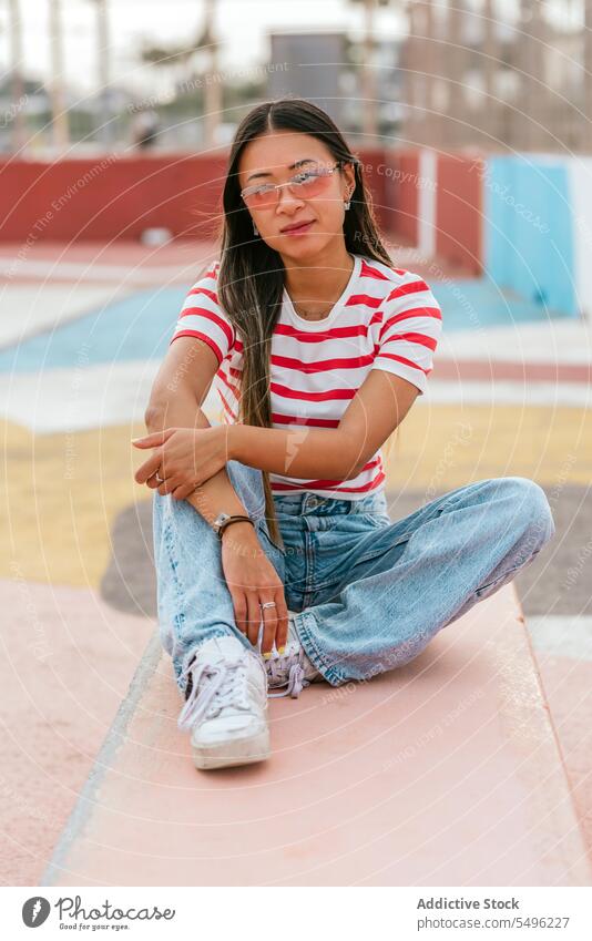 Young Asian woman sitting on stone bench dreamy thoughtful pensive calm alone casual eyewear eyeglasses peaceful trendy content attractive pleasant dark hair