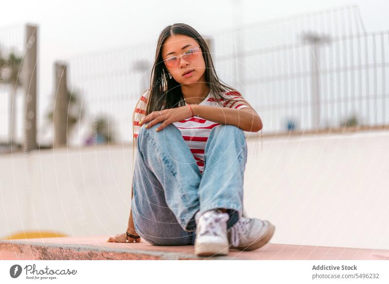 Young Asian woman sitting on stone bench dreamy thoughtful pensive calm alone casual eyewear eyeglasses peaceful trendy content attractive pleasant dark hair