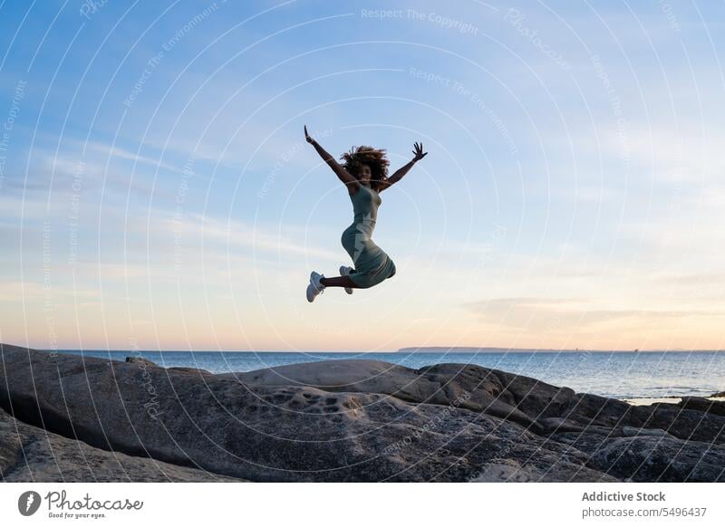 Happy African american young woman in dress looking at camera while jumping on rock against beach black lady female curly hair afro african american ethnic