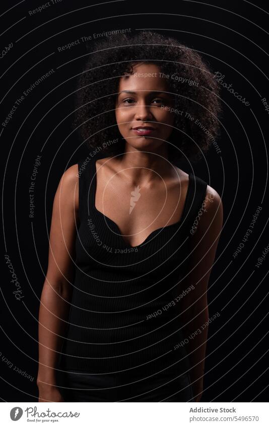 Smiling ethnic woman looking at camera in dark studio positive smile appearance leisure carefree happy afro curly hair friendly young female charming glad