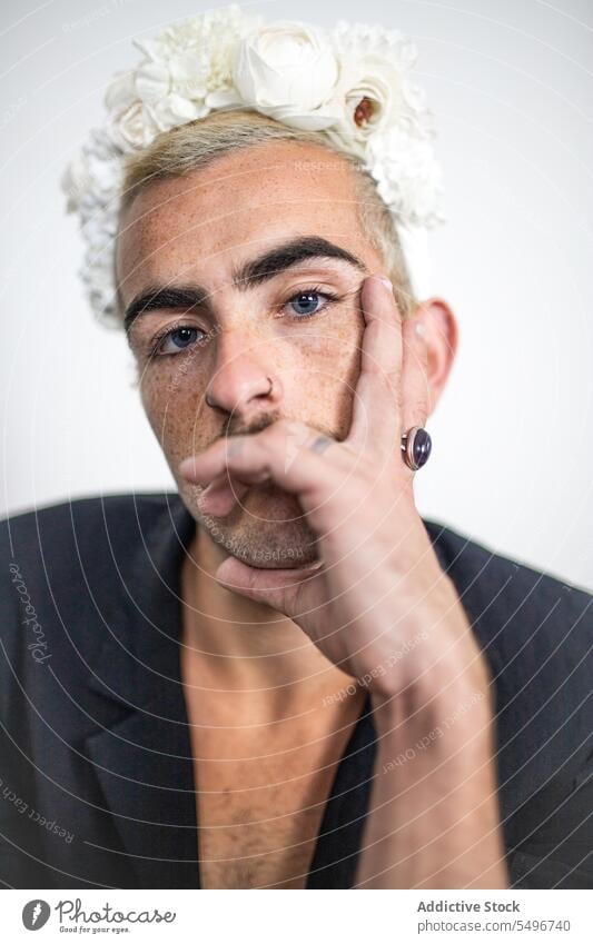 Confident young man with blue eyes and flower crown near white wall in room portrait calm makeup appearance unshaven mustache short hair confident male serious