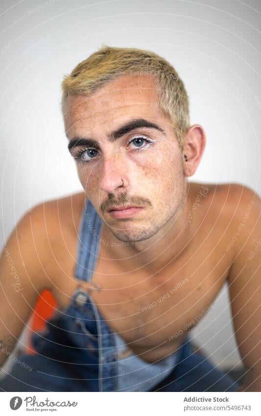 Thoughtful shirtless man sitting on chair near white wall in daylight makeup confident bare shoulders jeans palm lean forward male young individuality calm