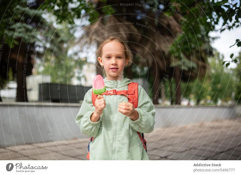 Girl with backpack standing near grassy field and enjoying ice cream child park path cute childhood green kid girl adorable summer sweet carefree wavy hair