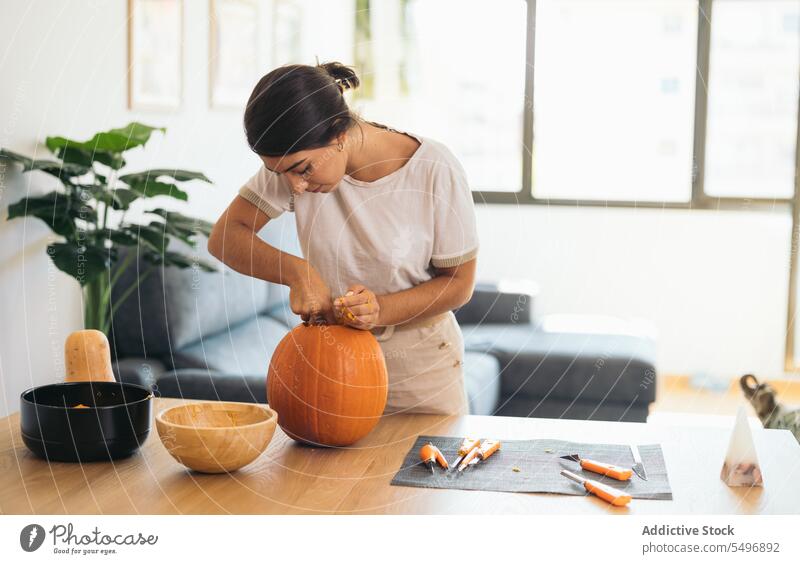 Concentrated young ethnic woman standing at table with pumpkin and Halloween tools halloween focus concentrate pulp pull serious at home female hispanic finger