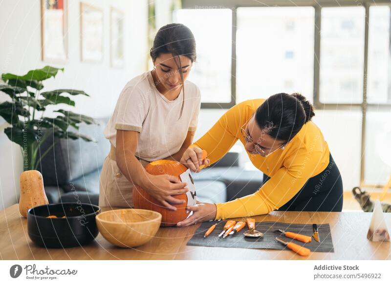 Young ethnic women standing at table with Halloween paper print on pumpkin friend jack o lantern halloween tool creative prepare at home female young hispanic