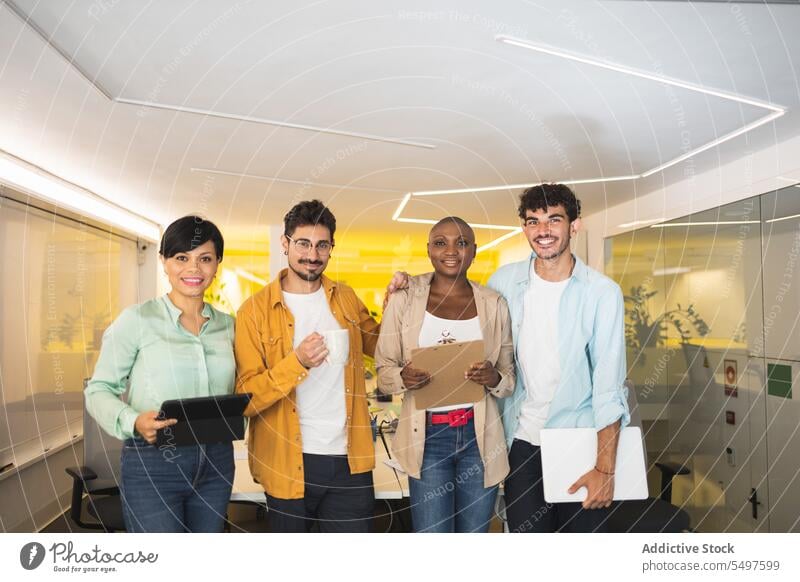 Happy multiethnic coworkers standing together in office colleague laptop workspace workplace tablet smile folder coffee group diverse multiracial cheerful team