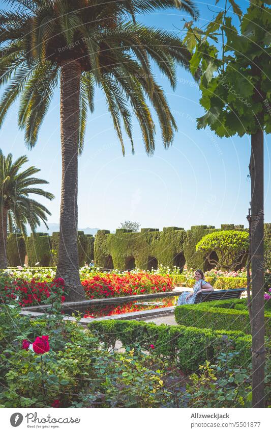 middle aged caucasian woman with blonde hair sits on a bench in a beautiful park next to a palm tree and a pool of water Summer Beautiful weather Cloudless sky