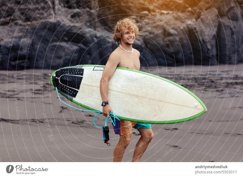 Fit young surfer man with curly blond hair with surfboard goes by the ocean having fun doing extreme water sports, surfing. Travel and healthy lifestyle concept. Sports travel destination.