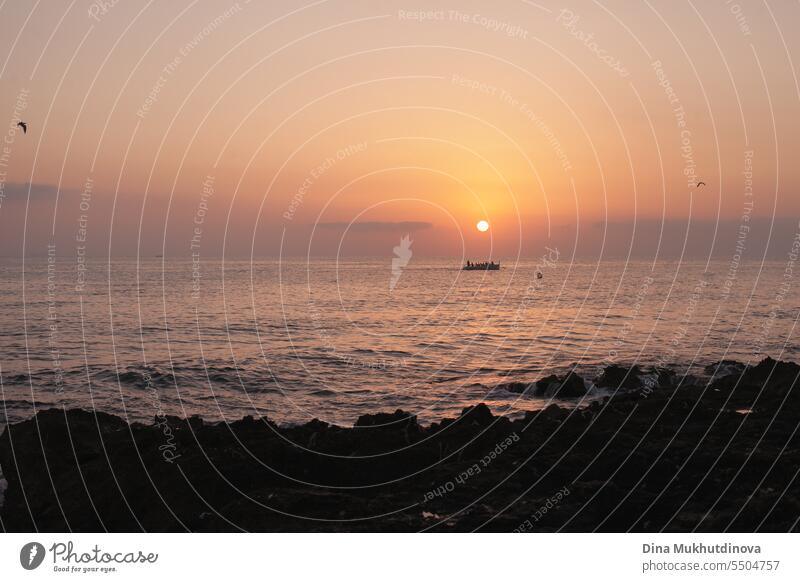 Rowing boat and birds at sunrise over the sea in early morning light. Sun rising over the water on rocky beach. Sunset landscape. Water splashing with waves.