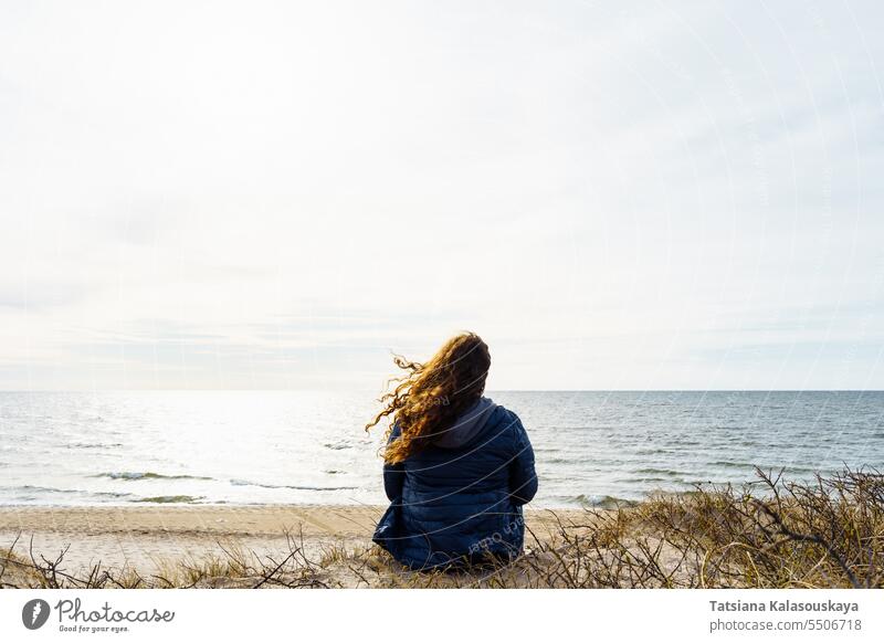 Curly-haired woman in a thin down jacket sitting on the seashore in the evening, hair fluttering in the wind plus size Baltic Sea nature beauty beach coastline