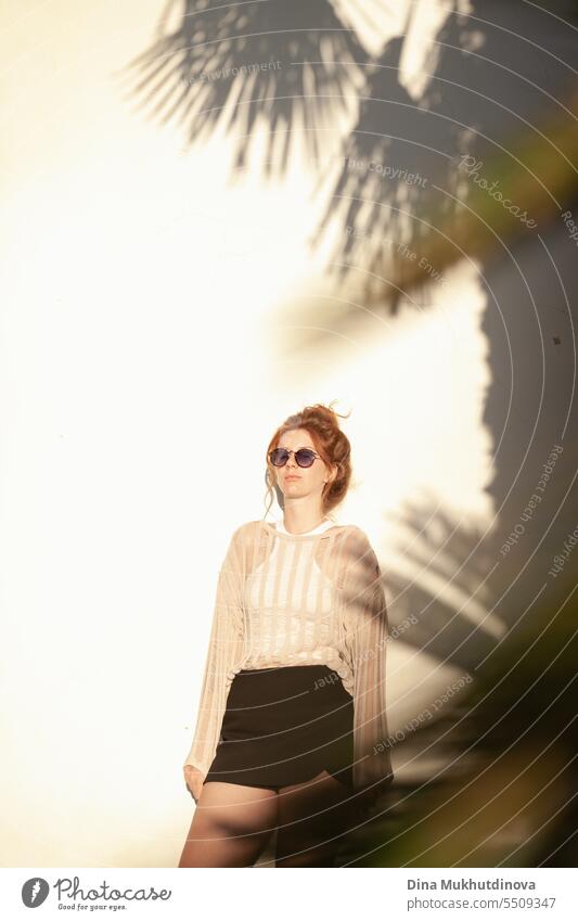 young beautiful red haired woman in casual clothes  in sunglasses with hair bun on beige wall background with palm tree shadows. Candid outdoor portrait in the park.