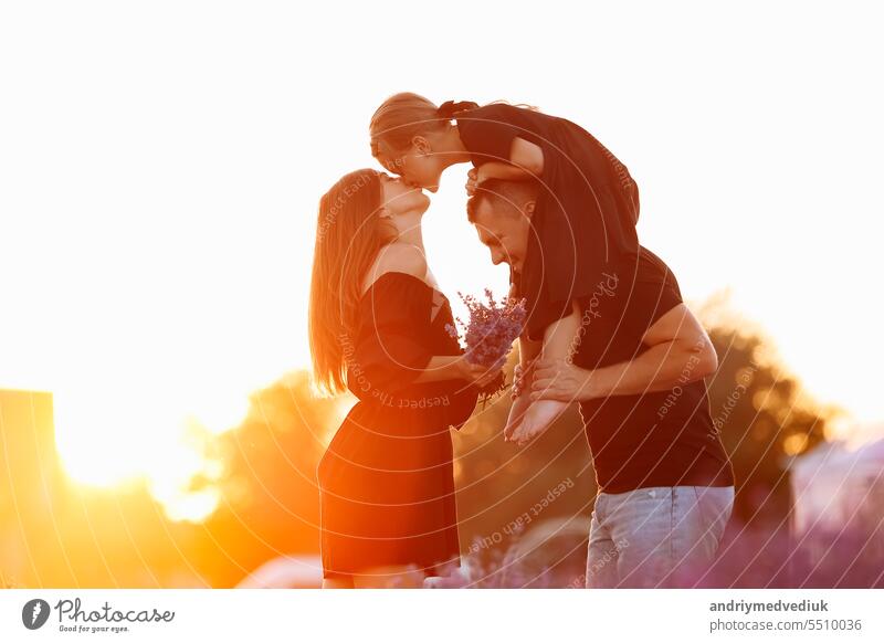 happy family day. Young mother and father carrying on shoulders daughter in lavender field on sunset. Dad, mom and child girl kissing and hugging on nature on summer. friendly family. family look