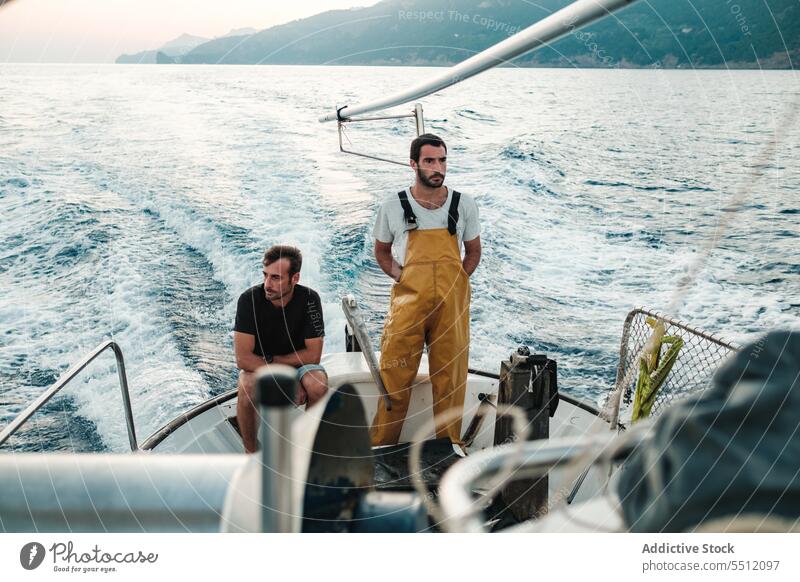 Young ethnic fishermen riding on fishing boat on rippling seawater fisherman travel sailboat mountain sky seascape ripple nature male young hispanic trip vessel