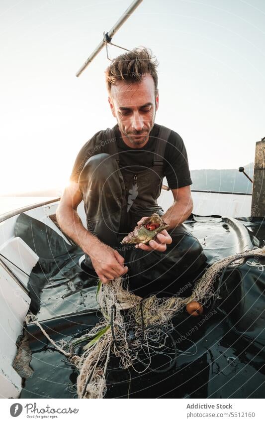 Concentrated young ethnic man sitting on deck with mollusk fishing net catch focus cloudless blue sky summer male hispanic fisherman daytime concentrate nature