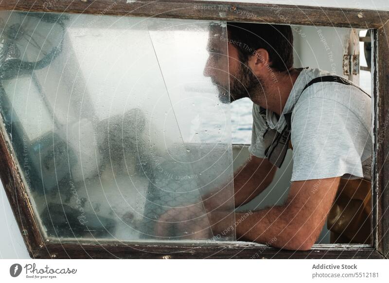 Bearded ethnic man in overalls sitting in boat cabin near dashboard sea window travel sail water transport summer male hispanic mallorca spain soller daytime