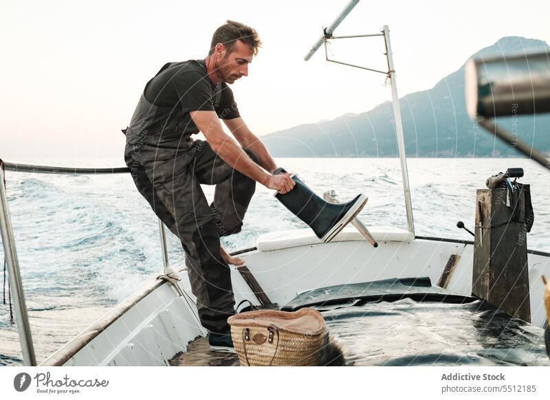 Young ethnic man standing on boat and wearing rubber sailing shoe sailboat sea evening railing seascape fence metal lean on seawater male young hispanic spain