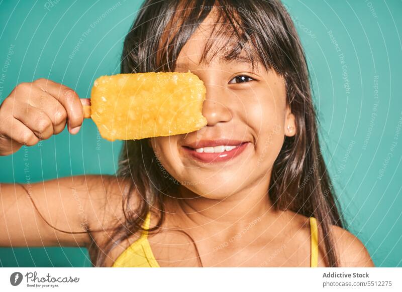 Happy ethnic girl child standing with popsicle near face against turquoise backdrop ice cream sweet cute delicious smile adorable kid asian colorful tasty