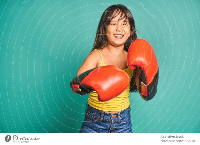 Cheerful ethnic girl child standing in boxing gloves near turquoise backdrop kid smile portrait glad positive cheerful room playful boxer appearance light