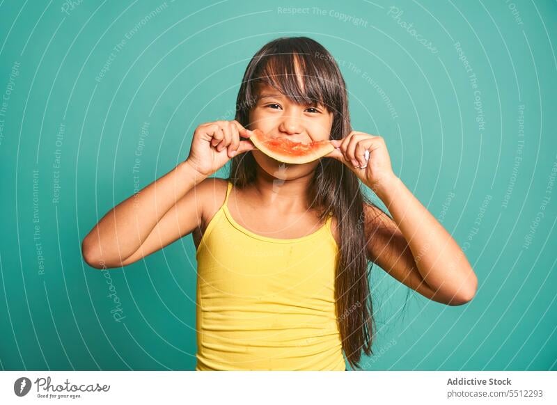 Happy ethnic girl child standing and eating piece of watermelon against turquoise backdrop kid delicious appearance sweet cute tasty adorable asian long hair