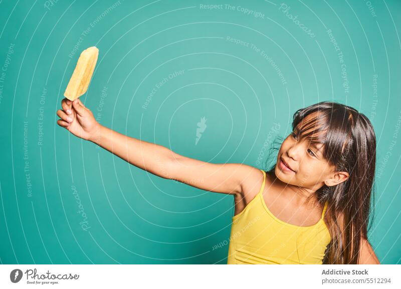 Happy ethnic girl child standing with popsicle in hand against turquoise backdrop kid childhood show frozen cute delicious happy playful asian black hair