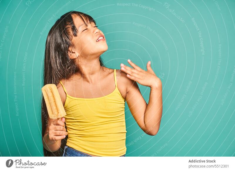 Tired ethnic girl child standing with popsicle in hand against turquoise backdrop kid eyes closed summer adorable cute wave hand carefree calm innocent asian