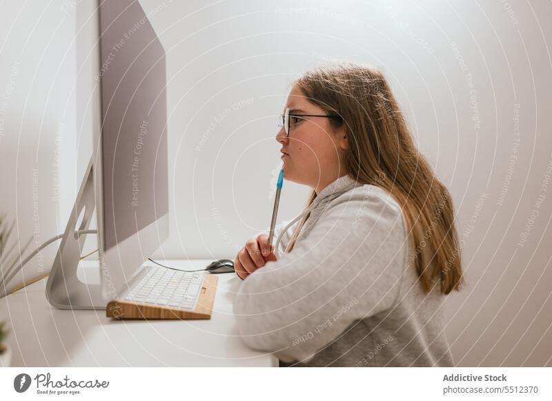 Serious girl sitting at table with pen and looking at monitor at home computer potted plant desk bright light teen teenage focus laptop gadget device internet