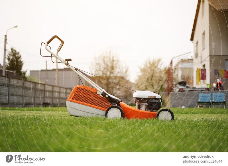 Modern orange-grey electric lawn mower on bright lush green lawn at residential backyard of house. Gardening work tools. Rotary lawn mower machine cut grass. Professional lawn care service.