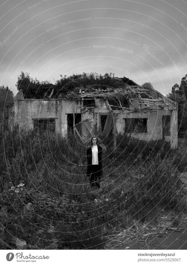 A young woman stands alone in a rugged setting, surrounded by tall grasses and an abandoned, crumbling building in the background portrait old house