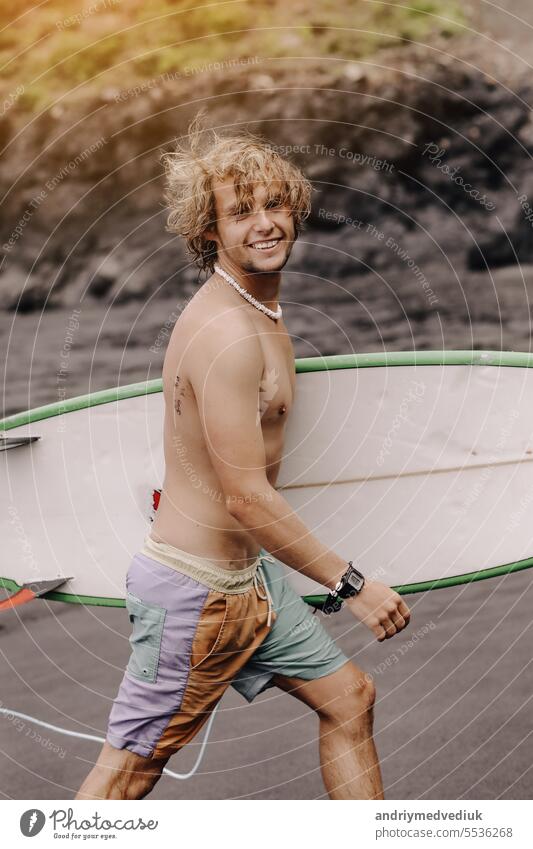 Handsome fit young blond man with mock up surfboard waits for wave to surf spot at sea ocean beach with black sand and looks at camera. Concept of sport, fitness, freedom, happiness, new modern life